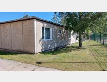 Ein einstöckiges Gebäude mit grauer Fassade und mehreren weißen Fenstern, umgeben von einer Rasenfläche. Links ein Gehweg, rechts ein Maschendrahtzaun und ein Baum, der teilweise Schatten spendet.