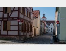 Schmale, gepflasterte Straße in einer Altstadt, flankiert von Fachwerkhäusern mit farbigen Fassaden und Holzfenstern. Im Hintergrund ist ein Kirchturm mit Uhr und Wetterfahne zu sehen.