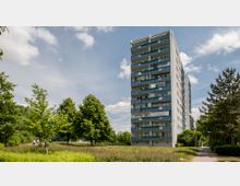 Ein Hochhaus mit mehreren Etagen und Balkonen steht am Rand einer grünen Parkanlage mit Bäumen und Wiesen, im Vordergrund verläuft ein gepflasterter Weg. Der Himmel ist teils bewölkt mit blauem Himmel im Hintergrund.