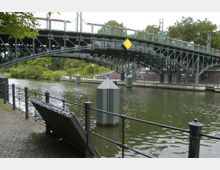 Stählerne Brücke über einen Fluss oder Kanal, umgeben von Grünflächen und Uferpromenaden mit Geländern; im Vordergrund ein Schild mit dem Schriftzug "Schulenburg".