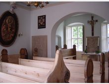 Innenraum einer kleinen Kapelle mit hölzernen Kirchenbänken, einem Altar mit Kerzen und religiösen Skulpturen. Große Fenster lassen Tageslicht herein, und an den Wänden hängen christliche Symbole und Dekorationen.