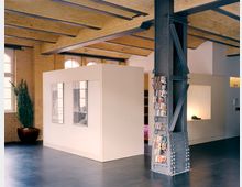 Moderner Innenraum eines Lofts mit gelben Backsteinwänden, freiliegenden Stahlträgern und einem hellbeigen freistehenden Raumteiler. Ein großes Fenster, eine industrielle Stütze mit Regalen für CDs und ein dunkler Holzboden prägen die Gestaltung.