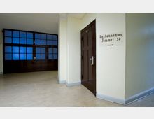 Flur in einem Gebäude mit beigem Linoleumboden und hellgelben Wänden. Rechts eine braune Tür mit der Nummer 57, daneben ein Schild mit der Aufschrift „Postannahme Zimmer 34“ und einem Pfeil; im Hintergrund eine Glastrennwand mit blauen Fensterscheiben und einer Tür.