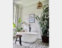 Modernes Badezimmer mit freistehender weißer Badewanne, schwarzer Armatur und gemusterten Fliesen. Pflanzen in Körben und ein hängender Lampenschirm aus Rattan ergänzen die dezente Einrichtung, während ein Bild an der Wand die Raumdekoration abrundet.