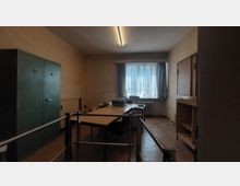 Ein schlicht eingerichteter Büroraum mit einem rechteckigen Tisch, mehreren Stühlen, einem großen grünen Schrank an der linken Wand und einem Fenster mit Vorhängen im Hintergrund. An der rechten Wand befinden sich Holzschränke, und auf dem Tisch stehen einige Bürogeräte.