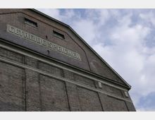 Ziegelfassade eines historischen Lagerhauses mit der Aufschrift „GETREIDE SPEICHER“ und kleinen, rechteckigen Fenstern unter einem Giebeldach, vor einem bewölkten Himmel.