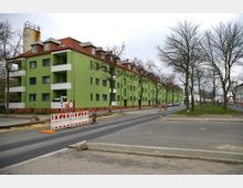 Mehrstöckiges Wohngebäude mit grün gestrichener Fassade und roten Ziegeldächern, an einer Straße mit Baustellenabsperrungen und kahlen Bäumen im Vordergrund. Die Umgebung wirkt urban, mit Gehwegen und vereinzelter Bebauung im Hintergrund.