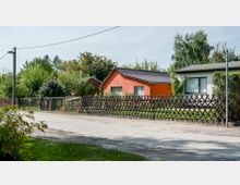 Eine ländliche Straße mit kleinen Häusern, darunter ein orangefarbenes Haus mit dunklem Dach und ein helles Haus mit Flachdach, umgeben von Gärten und einem Holzzaun. Im Hintergrund sind grüne Bäume und Sträucher zu sehen.