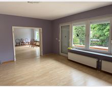 Wohnraum in einem Haus mit hellem Laminatboden und lila gestrichenen Wänden. Große Fenster mit Blick auf einen Garten und eine Terrasse, weiße Heizkörper unter den Fenstern; im angrenzenden Zimmer befinden sich Holzstühle, die entlang der Wand aufgestellt sind.