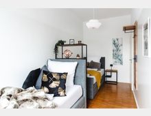 Modernes Schlafzimmer mit zwei grauen Einzelbetten, die durch ein Regal mit Deko-Elementen getrennt sind. Der Raum hat helle Wände, einen Holzboden, minimalistische Möbel und eine grüne Pflanzendekoration.