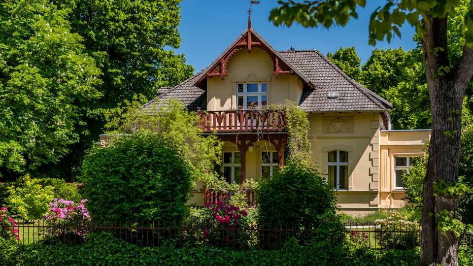 Ein zweigeschossiges, helles Landhaus mit Satteldach und Holzbalkon, umgeben von dichtem Grün, Hecken und blühenden Sträuchern hinter einem niedrigen Zaun. Links im Vordergrund steht ein gelbes Wegweiserschild am Straßenrand.