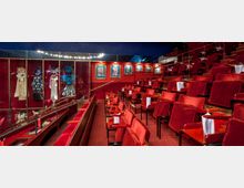 Sitzbereich eines Theaters mit roten Polstersesseln und kleinen Holzklapptischen, die Reihensitze terrassenartig anordnen. Im Hintergrund sind gläserne Vitrinen mit bunt dekorierten Bühnenkostümen und Bilderrahmen an einer roten Wand zu sehen.