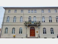 Fassade eines historischen Gebäudes mit beigen Wänden, weißen Fenstern im Rundbogenstil und einem großen Holzeingangstor. Über dem Eingang befindet sich ein kleiner Balkon mit Blumen und einem Wappen, flankiert von zwei Laternen.