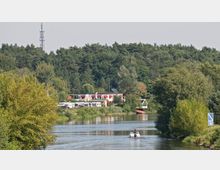 Ein Flusslauf in einer waldreichen Umgebung mit einem Freizeitgelände am Ufer, das aus einem Gebäude mit roten Markisen und mehreren parkenden Wohnmobilen besteht. Ein kleines Boot fährt auf dem Fluss, und im Hintergrund erstreckt sich ein dichter Wald.