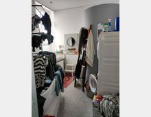 A cluttered laundry room with a tiled floor, featuring a washing machine, stacked dryer, and drying racks filled with clothes. The space also includes a curved wall, a small sink with a circular mirror, and various household items scattered throughout.