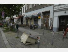 Straßenszene mit einem Imbiss namens "Kebabhaus-Grill bei Ismail" in einem alten Gebäude mit grauer Fassade und Graffiti. Vor dem Imbiss stehen Holztische und Bänke auf dem Gehweg, daneben befinden sich Bäume und parkende Fahrräder. Möbel liegen auf der Kopfsteinpflasterstraße, und Poller grenzen den Gehweg ab.