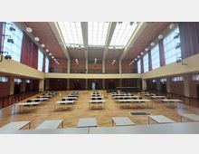 Großer Veranstaltungssaal mit Holzfußboden und einer hohen Decke, die mit großen Oberlichtern ausgestattet ist. Der Raum ist mit Reihen von Tischen und Stühlen eingerichtet, umgeben von hohen Fenstern mit roten Vorhängen und dekorativen Hängeleuchten.