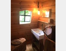 Kleines Badezimmer mit Holzwänden und rustikaler Einrichtung, einschließlich eines hölzernen Toilettensitzes, eines rechteckigen Waschbeckens mit einem einfachen Metallwasserhahn und eines kleinen Wandspiegels. Ein schräg eingesetztes Fenster mit Sprossen lässt Tageslicht herein, während eine hängende Glühbirne den Raum beleuchtet.