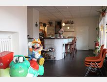 Café-Innenraum mit Bartresen aus hellen Fliesen, Holzbarhockern und Regalen mit Gläsern und Flaschen im Hintergrund. Im Vordergrund steht eine bunte Kinderspielfigur mit Comic-Motiv, daneben einige Stühle und Pflanzen.