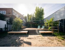 Außenbereich mit einer Holzbühne, die von kleinen Bäumen und Gerüsten umgeben ist. Links ein Backsteingebäude mit Graffitis, rechts Container, im Hintergrund Sonnensegel und Sitzmöbel.