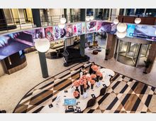 Modernes Hotellobby-Interieur mit einer geschwungenen, weißen Rezeption, dekoriert mit rosa Blumen in Vasen. Im Hintergrund eine Wendeltreppe, Sitzbereiche und eine gläserne Drehtür, umgeben von hellen Bodenmustern und einem umlaufenden LED-Bildschirm mit bunten Illustrationen.