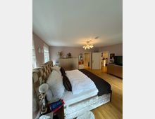 Ein großes Schlafzimmer mit Holzboden und hellvioletten Wänden. Im Vordergrund steht ein Bett mit gesteppter Kopfstütze, weißer Bettwäsche und dunklem Überwurf; im Hintergrund befinden sich eine Kommode, ein offener Durchgang und eine Tür.