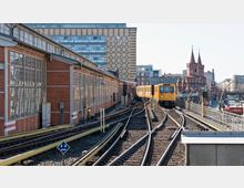 Eine gelbe U-Bahn fährt auf oberirdischen Gleisen neben einem Backsteingebäude mit großen Fenstern. Im Hintergrund sind moderne und historische Gebäude, darunter die markanten Türme der Oberbaumbrücke, zu sehen.
