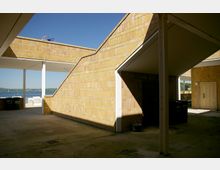 Ein moderner Außenbereich mit gelben Backsteinwänden und schrägen architektonischen Elementen. Im Hintergrund ist ein See mit Segelbooten und eine Landschaft unter einem klaren blauen Himmel sichtbar.