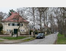 Straßenszene in einem Wohngebiet mit Kopfsteinpflaster, gesäumt von kahlen Birkenbäumen. Auf der linken Seite steht ein großes Haus mit hellgelber Fassade, grünem Tor und rotem Ziegeldach, daneben einige parkende Autos.