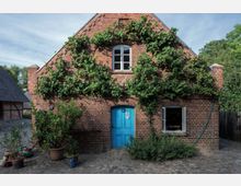 Ziegelstein-Gebäude mit Satteldach, an dessen Fassade Kletterpflanzen wachsen. Eine blaue Tür und ein Fenster mit weißen Rahmen sind von Pflanzen umgeben; vor dem Gebäude stehen Topfpflanzen auf einem gepflasterten Hof.