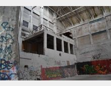 Ehemaliges Industriegebäude mit hohen Wänden und freiliegender Stahlkonstruktion an der Decke. Die Wände sind mit Graffiti bedeckt, und es gibt eine Betonstruktur mit offenen Räumen und Rostgeländern im Vordergrund.