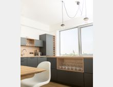 Moderne Küche mit grauen Schränken, Holzarbeitsflächen und offenem Stauraum für Gläser. Große Fenster lassen Tageslicht herein, während zwei hängende Lampen von der Decke herabreichen.