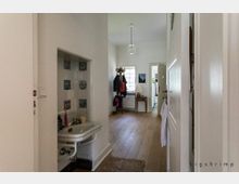 Flur in einem Wohnhaus mit hell gestrichenen Wänden, Holzboden und einer Tür im Vordergrund. Links ist ein kleines Waschbecken mit dekorativen Kacheln, rechts ein Garderobenständer mit Jacken und Taschen, im Hintergrund ein Fenster und ein Tisch mit Lampe.