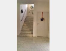 Treppenhaus mit weißen Wänden und Marmorboden; links führt eine Treppe mit Metallgeländer nach oben, rechts befindet sich eine weiße Tür mit einer sternförmigen Dekoration. Oben im Treppenbereich sind hängende Pflanzen und ein Fenster sichtbar.