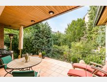 Überdachte Terrasse mit Holzdach, Terrakottafliesenboden und Blick auf einen üppig bewachsenen Garten mit Bäumen und Sträuchern. Die Terrasse ist mit einem Esstisch, grünen Stühlen, roten Liegestühlen, einem Heizstrahler und Pflanzen dekoriert.
