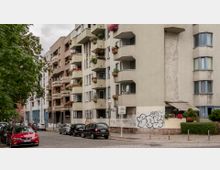 Mehrstöckiges Wohnhaus in einem städtischen Wohngebiet mit Balkonen, die mit Blumen und Pflanzen dekoriert sind. Im Vordergrund verläuft eine mit Kopfsteinpflaster bedeckte Straße mit parkenden Autos, und ein Baum säumt den Gehweg auf der linken Seite.