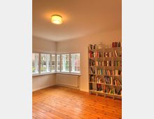 Heller Raum mit Holzdielenboden, bodentiefen Fenstern mit weißem Rahmen und einer Heizkörperleiste darunter. An der rechten Wand steht ein großes Regal, gefüllt mit Büchern und Schachteln.