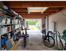 Innenansicht einer Garage mit sichtbaren Holzbalken an der Decke. Links befinden sich Regale, die mit Werkzeugen, Behältern und Materialien gefüllt sind, während rechts ein Fahrrad, Kabel und eine Sackkarre zu sehen sind. Im Hintergrund ist das geöffnete Garagentor mit Blick auf einen Gartenweg sichtbar.