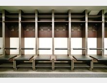 Reihe von Umkleidespinden in einem modernen Umkleideraum, mit dunklen Holzfächern, Haken und schwarzen gepolsterten Sitzen aus Metallrahmen. Der Boden ist mit Teppich ausgelegt, und die Beleuchtung ist hell.