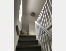 Treppenhaus mit grauem Teppichboden und weiß gestrichenen Geländern. Im oberen Bereich des Bildes sind eine Zimmertür, ein rundes Wanddekor und eine Deckenlampe zu sehen.