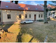 Ein beige gestrichenes Gebäude mit rotem Ziegeldach und mehreren rechteckigen Fenstern, umgeben von einer Rasenfläche mit vereinzelten Bäumen. Vor dem Gebäude steht ein weißer Oldtimer, und im Hintergrund sind weitere Gebäude sichtbar.