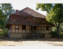 Fachwerkhaus mit bröckelndem Putz und einem Schindeldach, das teilweise verwittert ist, umgeben von Bäumen und naturbelassenem Gelände; eine überdachte Holztreppe führt zum Eingang.