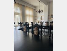 Ein Konferenzraum mit dunklen Holztischen und hochlehnigen, gepolsterten Stühlen mit Verzierungsmustern. Im Hintergrund befinden sich große Fenster mit vertikalen Jalousien, an den Wänden hängen Bilder, und von der Decke hängt ein schwarzer Kronleuchter.