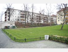 Grüne Parkfläche mit gepflegtem Rasen, umgeben von pflasterierten Wegen mit Metallgeländern. Im Hintergrund sind mehrstöckige Wohngebäude mit Balkonen sowie einige kahle Bäume zu sehen.