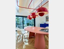 Moderner Besprechungsraum mit einem ovalen, pinkfarbenen Tisch, weißen Stühlen mit Holzbeinen und roten Hängelampen. Im Hintergrund große Fenster mit Blick auf grüne Vegetation und ein blaues Sideboard.