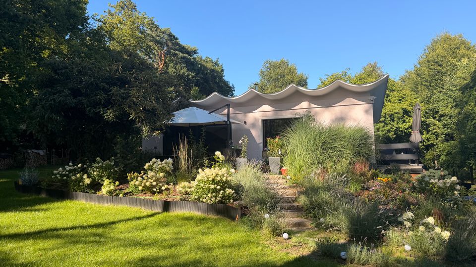 Modernes, flaches Gartenhaus mit markant geschwungenem Wellendach, umgeben von dichtem Baumbestand. Im Vordergrund liegen ein gepflegter Rasen, blühende Beete mit Ziergräsern und ein kleiner Treppenweg zur Terrasse mit Sonnenschirm.
