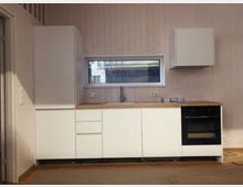 Moderne, minimalistische Küchenzeile mit weißen Schrankfronten und Holzarbeitsplatte vor hell vertäfelter Wand. Unter einem schmalen Fenster befinden sich Spüle und Armatur; rechts ist ein eingebauter Backofen, links ein hoher Hochschrank und darüber ein Hängeschrank.