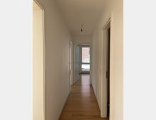 Flur eines Wohngebäudes mit hellen Wänden, Holzboden und mehreren Türöffnungen auf beiden Seiten. Am Ende des Flurs ist eine geöffnete Tür zu sehen, die zu einem Raum mit einem Fenster führt.