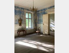 Ein Raum mit abgenutzten blauen Wänden, einem großen Holzfenster mit Blick nach draußen, Holzdielenboden, einem Kronleuchter an der Decke und antiken Möbelstücken, darunter ein kleiner verzierter Tisch mit Vase und ein einzelner Stuhl. Auf der rechten Seite steht eine weiße Holztür, die leicht geöffnet ist.