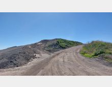 Unbefestigter Weg, der an einem Hügel entlangführt, mit einer Mischung aus kahlem Erdreich und vereinzeltem Grünbewuchs unter einem klaren blauen Himmel. Auf der linken Seite sind steinige Hänge, während auf der rechten Seite Gras die Erdoberfläche bedeckt.